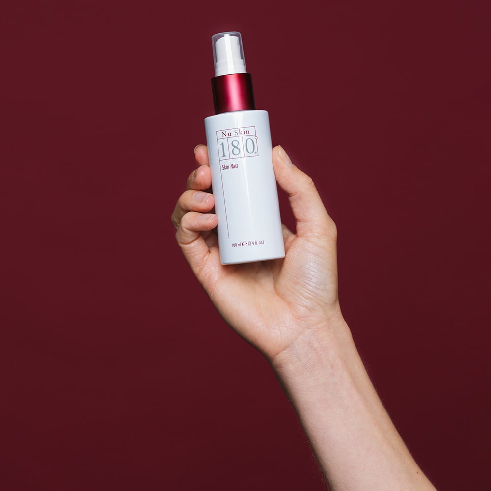 Hand hält 180° Skin Mist von NuSkin (geschlossen) vor weinrotem Hintergrund.