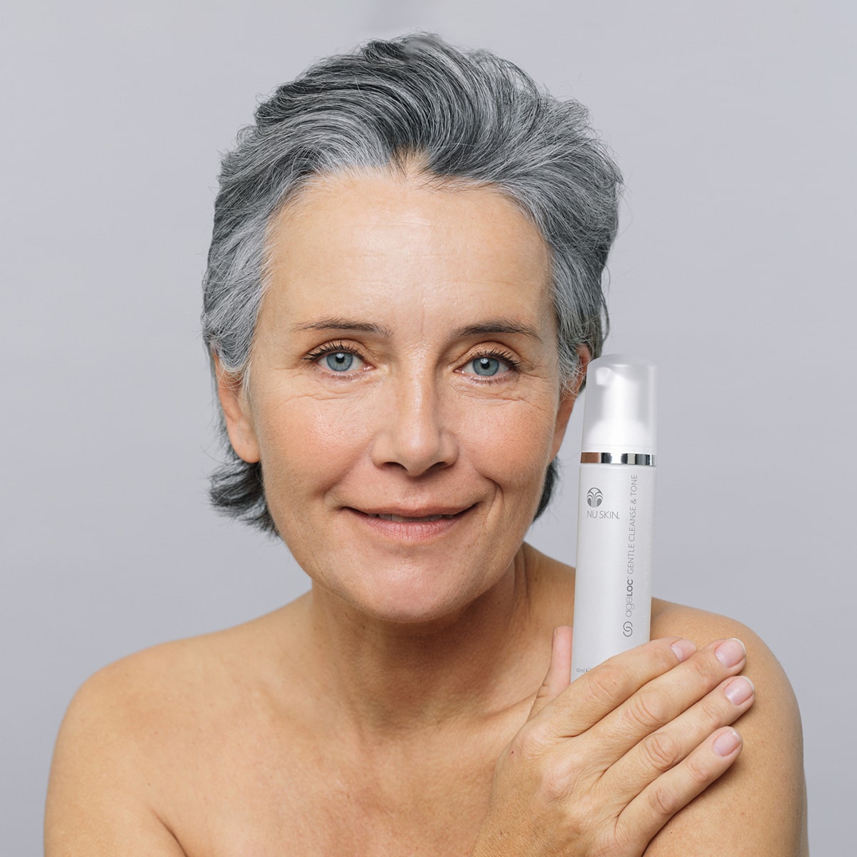 Frau mittleren Alters mit grauer Kurzhaarfrisur hält ageLOC Gentle Cleanse & Tone Dosierspender lächelnd neben ihr Gesicht.
