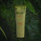 Epoch Ava Puhi Moni Shampoo and Light Conditioner von Nu Skin liegt geschlossen auf dunkelgrünen Farnblättern. Auf den Blättern und der Tube glänzen Wassertropfen.