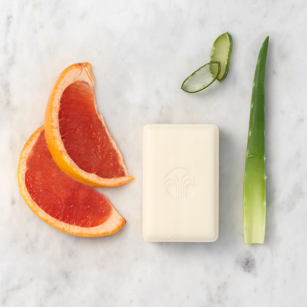 Body Bar liegt ausgepackt auf marmorner Oberfläche, daneben zwei aufgeschnittene Grapefruit-Scheiben und ein paar Blätter der Aloe Vera Pflanze.