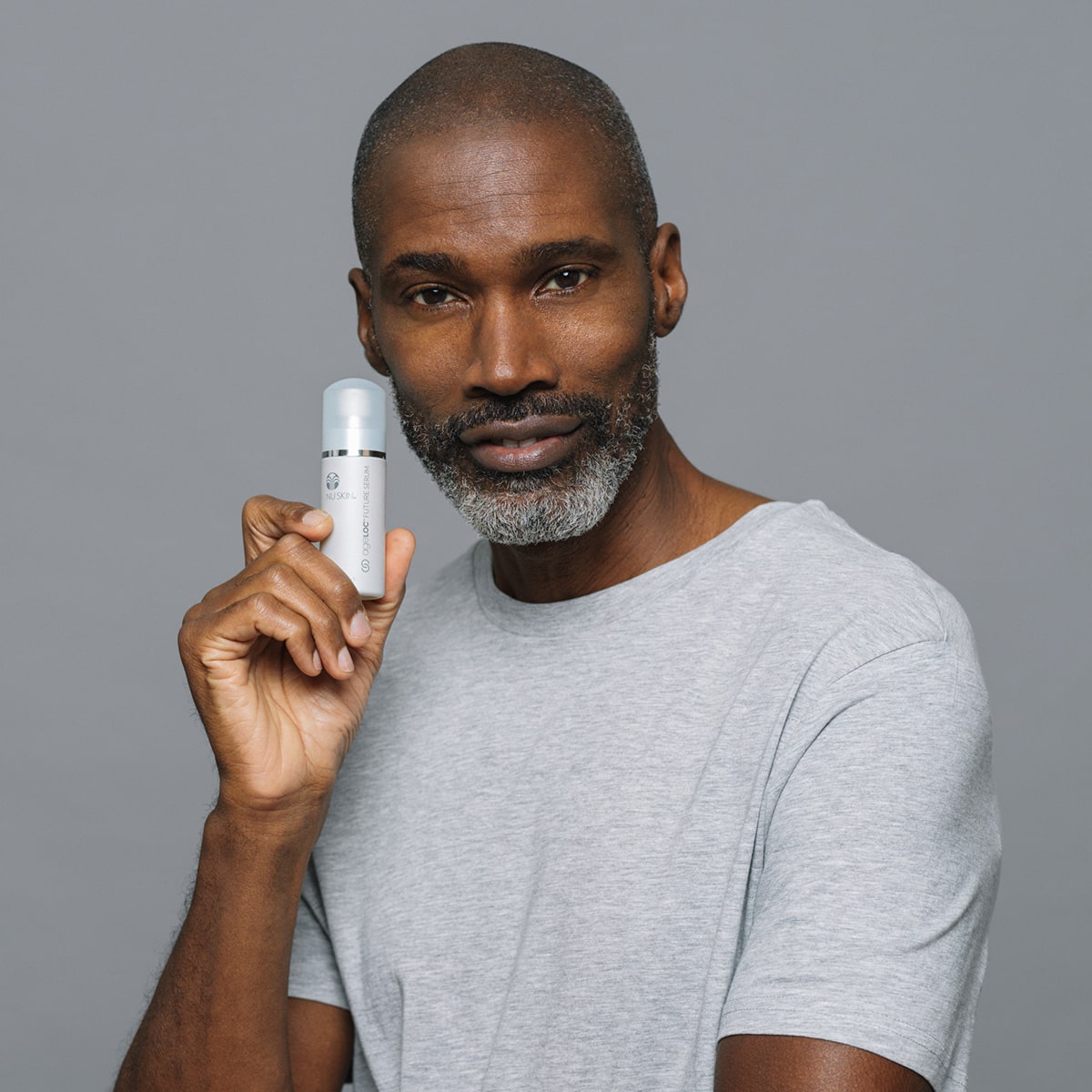 Mann mittleren Alters mit gepflegtem Bart und dunklem Teint hält lächelnd das ageLOC® Future Serum (30ml) in der Hand. Er steht vor einem grauen Hintergrund.
