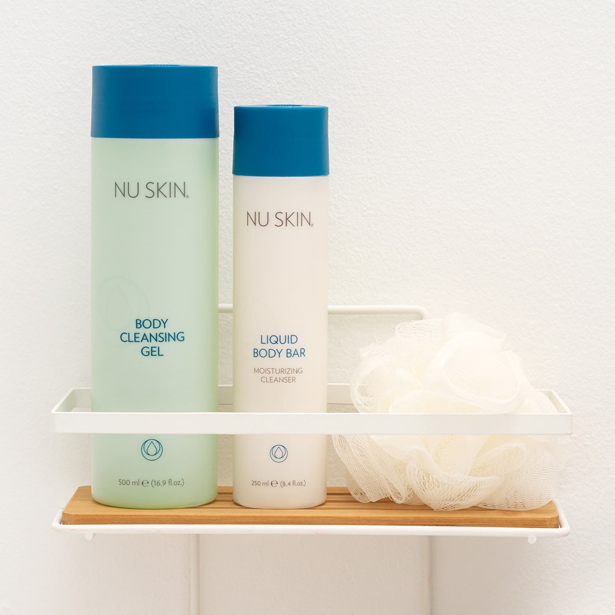 Nu Skin Basic Produkte (Body Cleansing Gel, 500ml und Liquid Body Bar, 250ml) stehen geschlossen auf einer kleinen Ablage in sanitärer Umgebung. Daneben liegt ein weißer Schwamm.