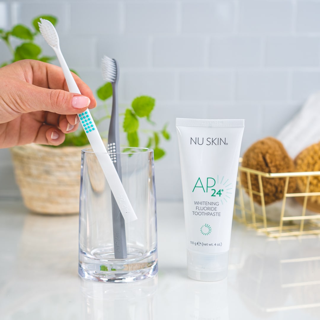 NuSkin AP 24 Whitening Fluoride Zahnpasta steht in sanitärer Umgebung neben Glas mit zwei Zahnbürsten. Hand greift nach einer von den beiden Bürsten.