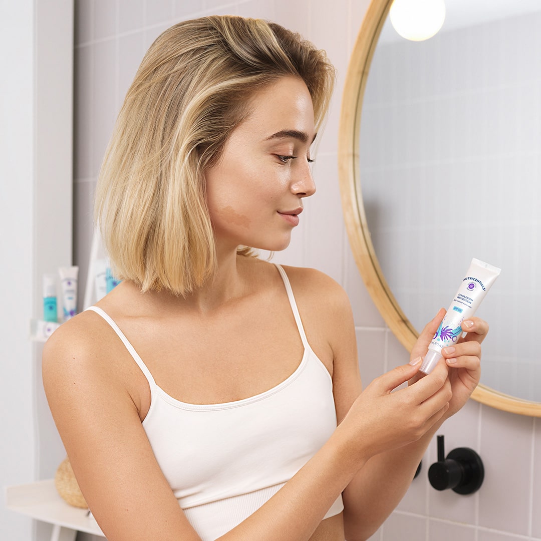 Junge Frau mit blonden Haaren steht lächelnd vor einem Badezimmerspiegel. Sie hält die Complexion Protection Daily Mineral Sunscreen von Nutricentials in den Händen.