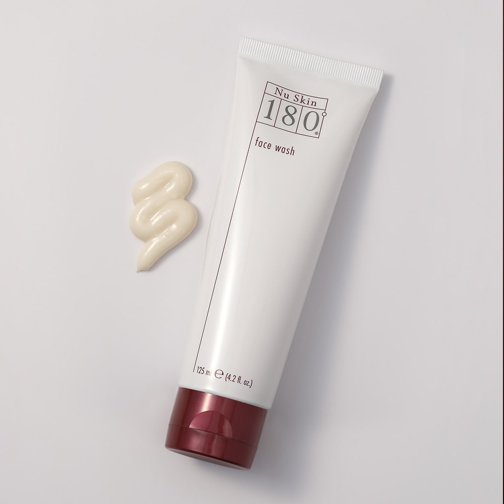 Nu Skin 180° Face Wash Tube (125ml) liegt geschlossen auf glatter weißer Oberfläche. Daneben wurde eine kleine Konsistenzprobe des cremigen, cremefarbenen cleansers aufgetragen.