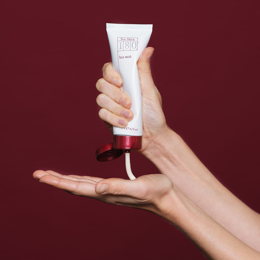 NuSkin 180° Face Wash wird aus geöffneter Tube vor weinrotem Hintergrund auf Handfläche gegeben.