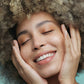 Junge Frau mit hellem Afro und frechen Sommersprossen hält sich lachend die Hände an das glatte Gesicht.