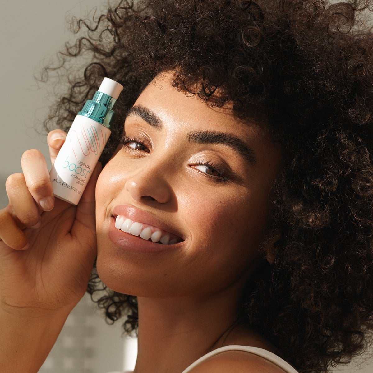 Junge Frau mit strahlendem Teint und wilden dunklen Locken hält das ageLOC Boost Activating Serum lächelnd an ihr sonniges Gesicht.