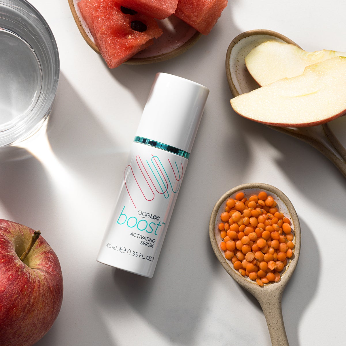 ageLOC Boost Activating Serum von Nu Skin liegt geschlossen auf weißer Oberfläche. Der Spender ist umgeben von verschiedenen Zutaten wie Apfelscheiben und aufgeschnittener Wassermelone.