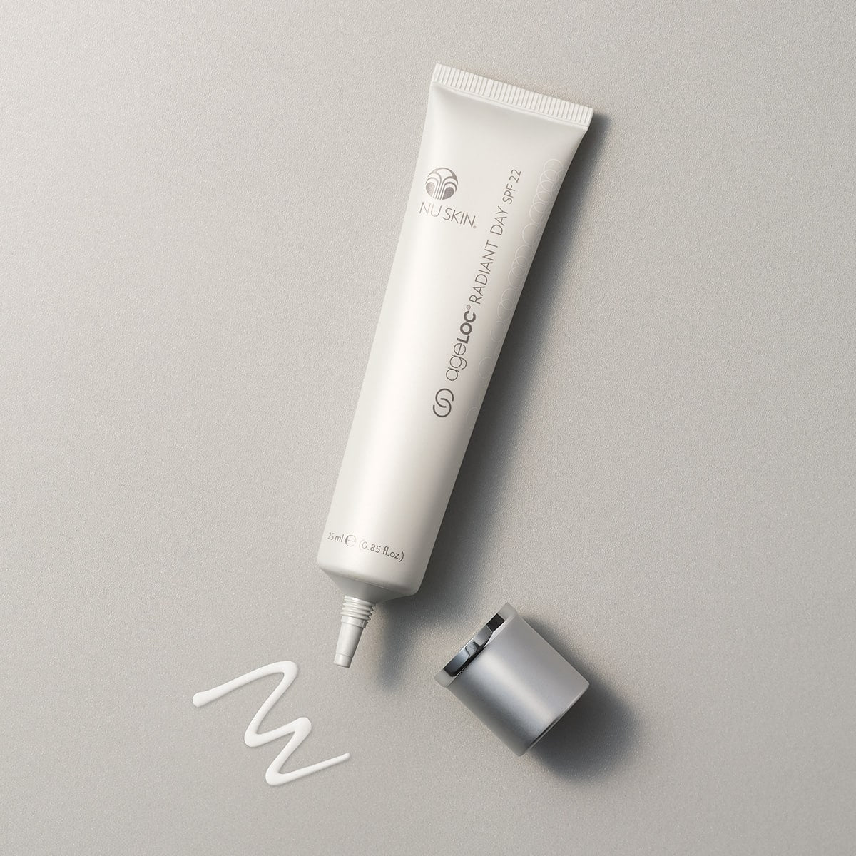 NuSkin ageLOC Radiant Day SPF 22 Tube liegt geöffnet auf glatter hellgrauer Oberfläche. Eine kleine Probe der weißen seidigen Lotion wurde vor der Tube aufgetragen.