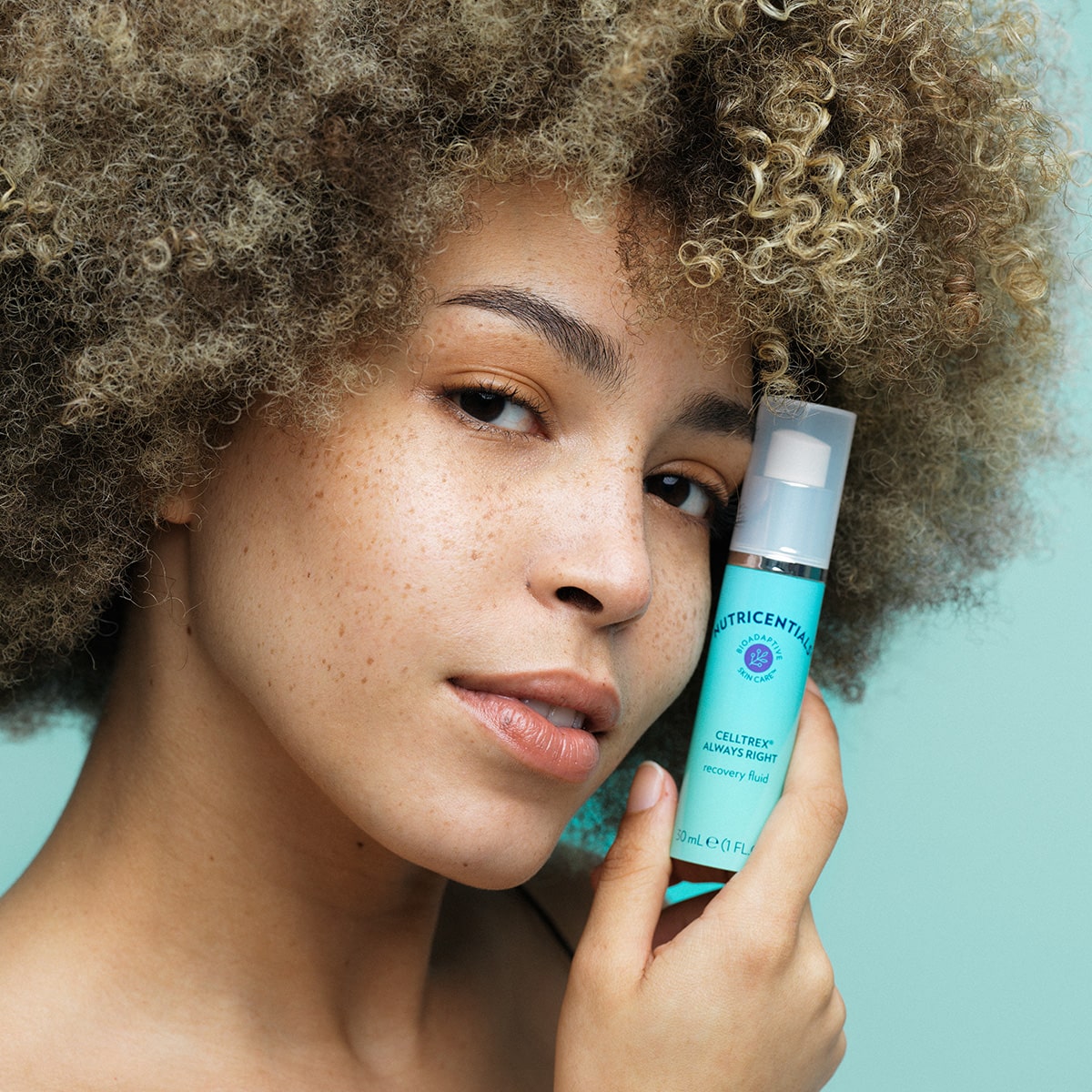 Junge Frau mit dunkelblondem Afro, Sommersprossen und glatter Haut hält die Celltrex Always Right Recovery Fluid neben ihr Gesicht.