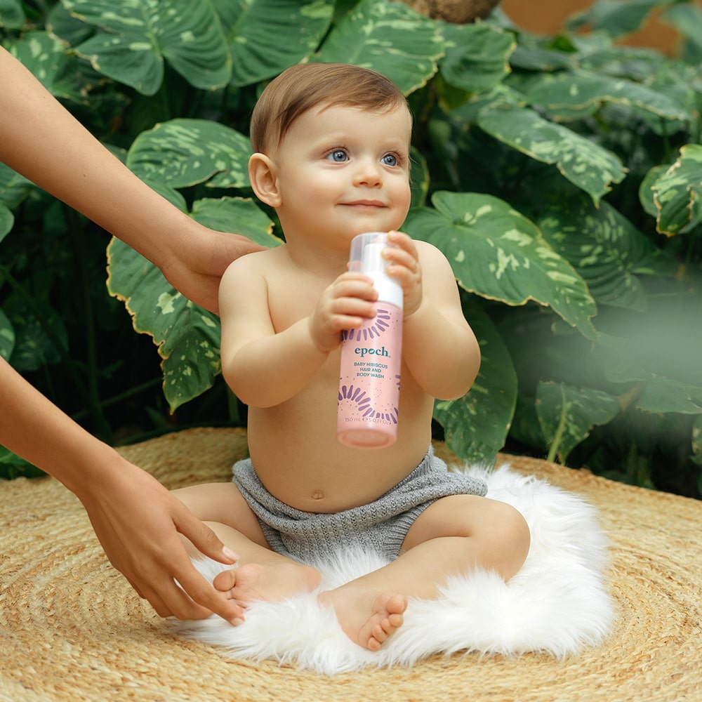Baby sitzt lächelnd vor dunkelgrünen Pflanzenblättern auf flauschigem Kissen. Es hält die Epoch Baby Hibiscus Hair and Body Wash Flasche von NuSkin in den Händen.