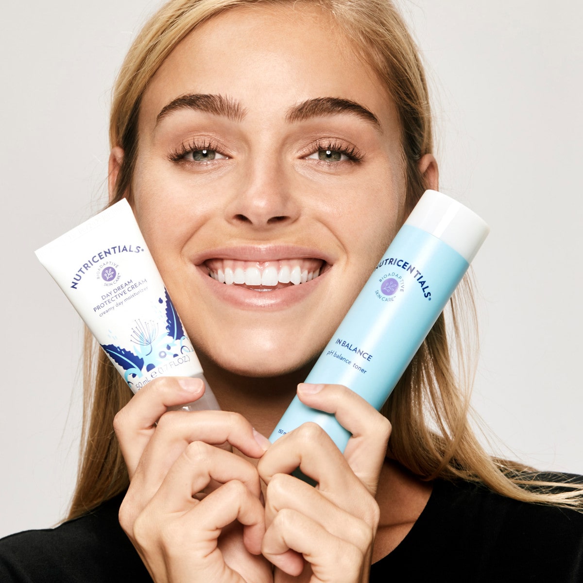 Junge Frau mit blonden Haaren und strahlendem Lächeln hält sich den In Balance pH Balance Toner und die Daydream Protective Cream (Nutricentials Produkte) an ihr Gesicht.
