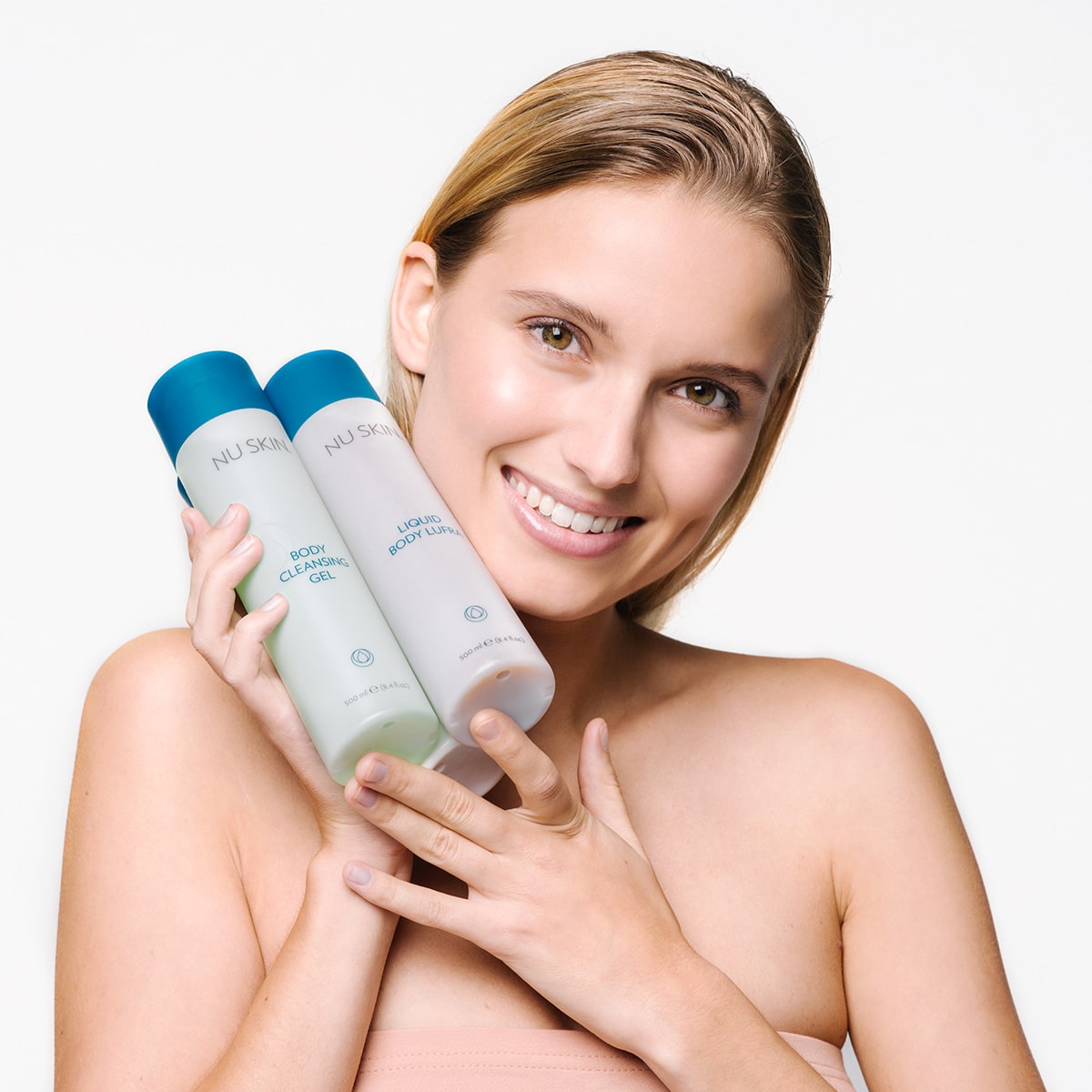 Junge Frau mit blonden frisch gewaschenen Haaren hält lächelnd Nu Skin Basic Produkte in den Händen: Body Cleansing Gel und Liquid Body Lufra.