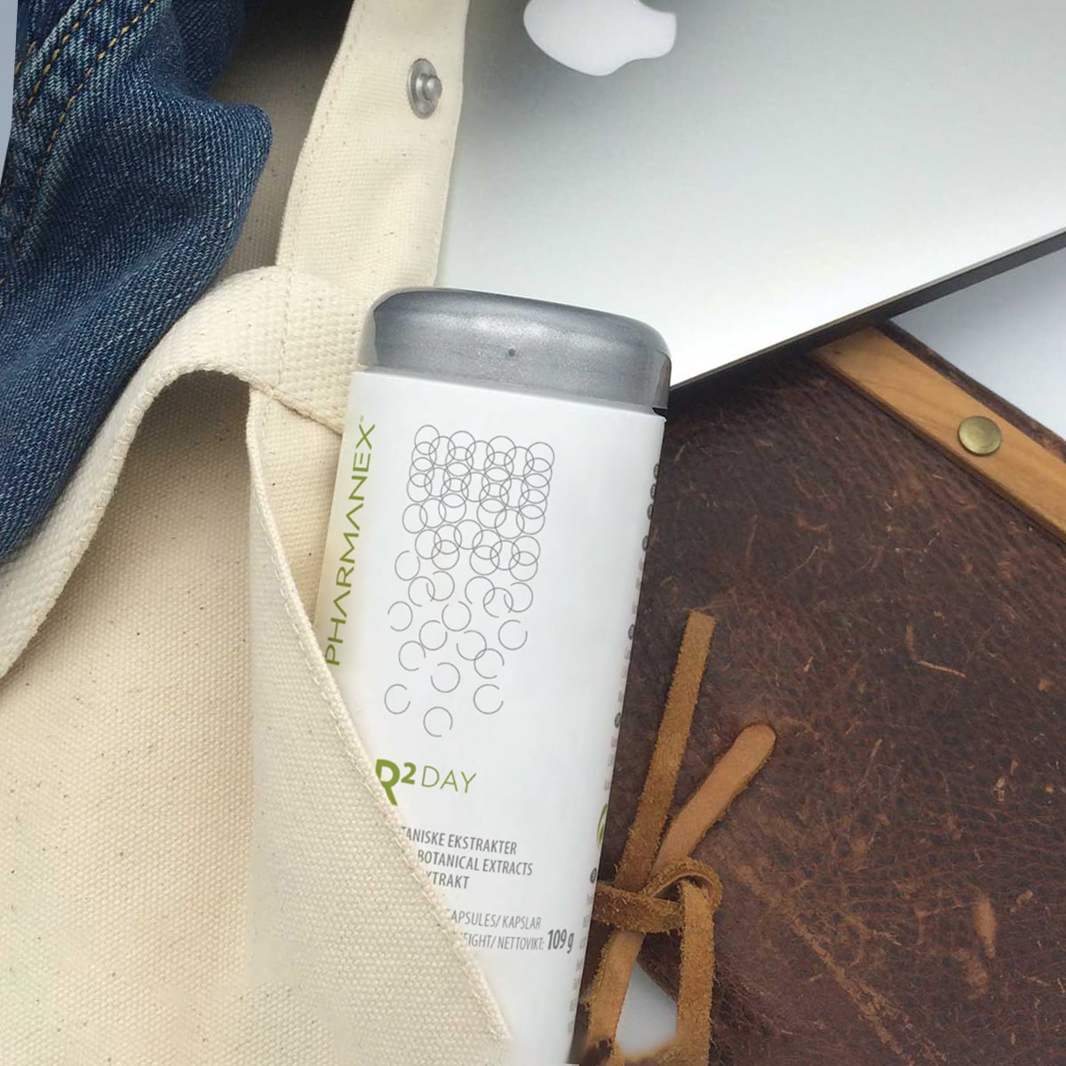 Pharmanex R² Day Dose ragt aus heller Stofftasche neben einem Notizbuch und Laptop hervor.