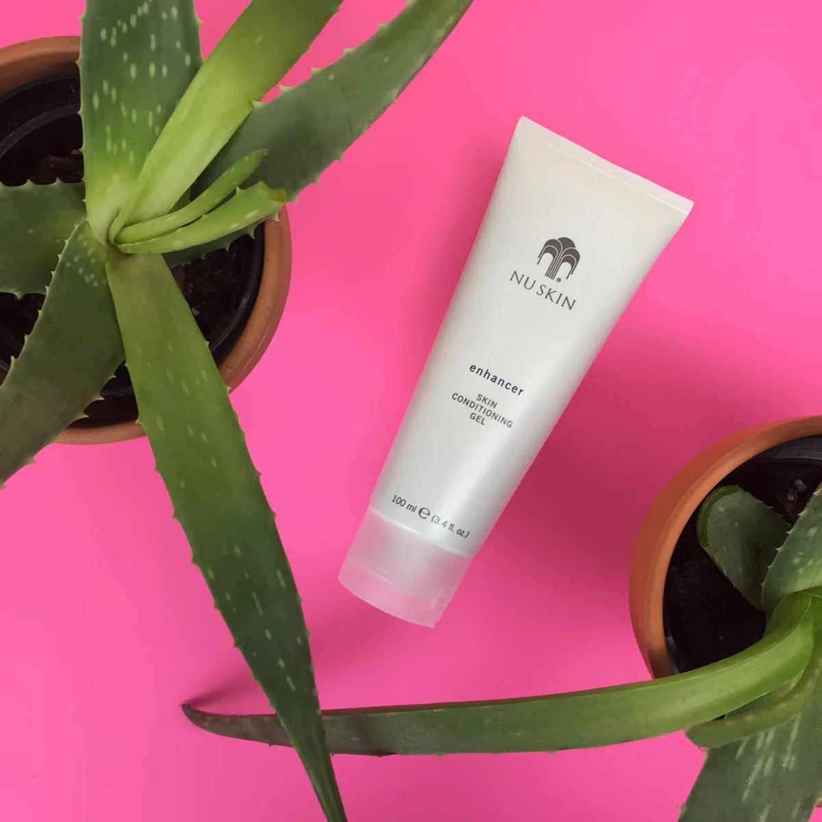 Enhancer Skin Conditioning Gel Tube (100ml) von NuSkin liegt auf rosa glatter Oberfläche zwischen zwei Aloe Vera-Blumentöpfen.