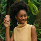 Junge Frau mit glattem dunklen Teint und Afrofrisur hält vor dunkelgrünem Farn lachend ein Stück NuSkin Epoch Polishing Bar.