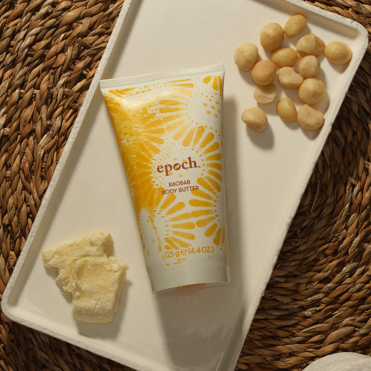 epoch Baobab Body Butter von Nu Skin liegt geschlossen auf einem kleinen weißen Tablett. Daneben eine handvoll Macadamia-Nüsse und ein paar Stücke Sheabutter.