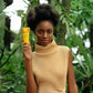 Junge Frau mit dunkler Haut und Afro steht in grüner Umgebung. Sie hält die epoch Firewalker Foot Cream von NuSkin lächelnd in der Hand.