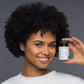 Junge Frau mit dunkler Afrofrisur und klaren braunen Augen hält lächelnd Eye Formular Glas von Pharmanex neben ihr Gesicht.