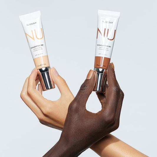 Die Hände zweier junger Frauen (eine mit schwarzem, die andere mit weißem Teint) halten passende Nu Colour Bioadaptive BB+ Skin Loving Foundation Tuben vor weißen Hintergrund.
