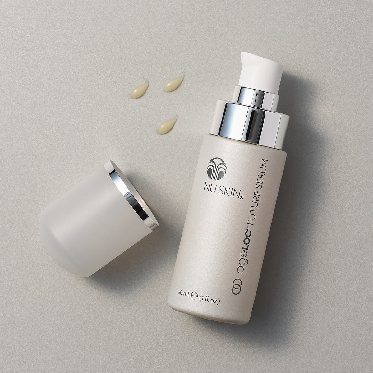 ageLOC® Future Serum Dosierspender (je 30ml) liegt geöffnet auf grauer Oberfläche. Neben der Packung wurden drei kleine Tropfen der Hautpflege aufgetragen.