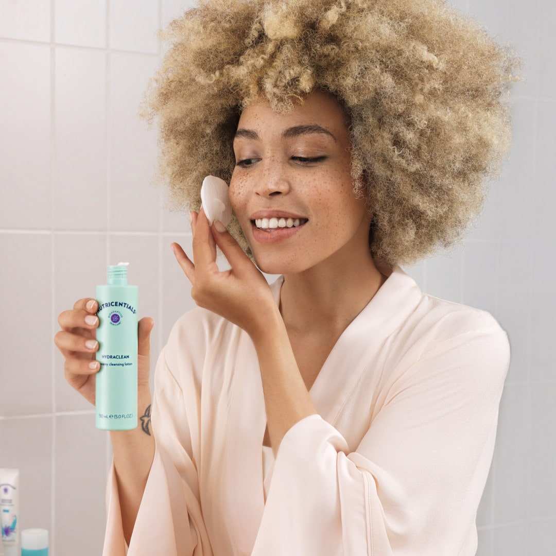 Junge Frau mit dunkelblondem Afro und glatter Haut tupft sich im Badezimmer mit einem Wattepad die Nutricentials Lotion auf die Wange.