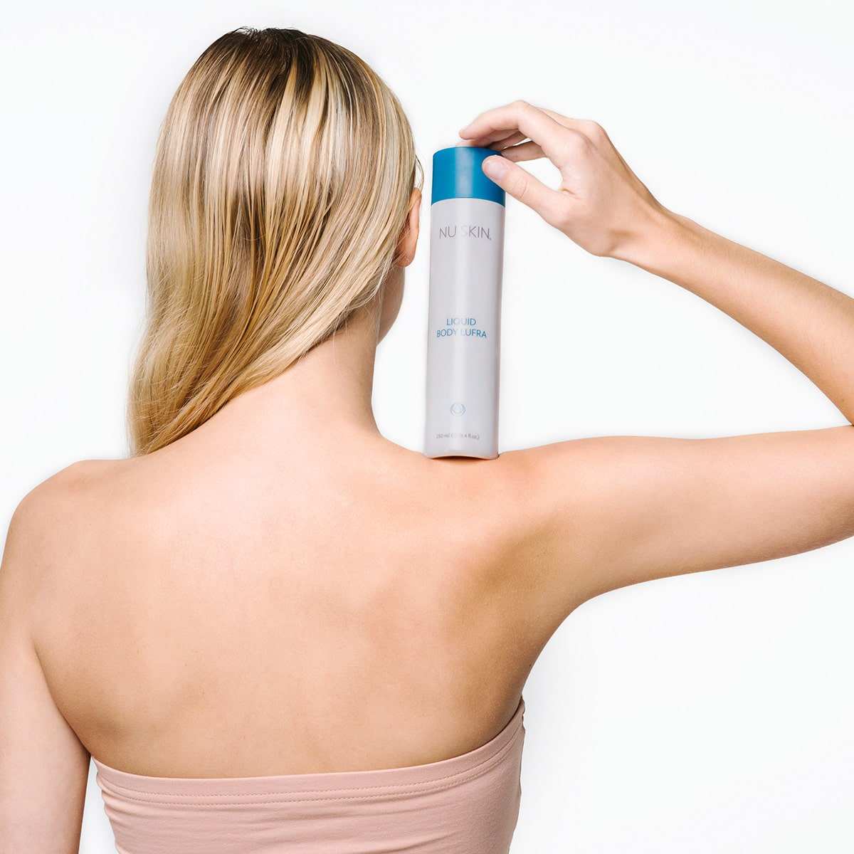 Junge Frau mit blondem glänzenden Haar steht den Rücken zu der Kamera gewandt. Mit der rechten Hand hat sie Liquid Body Lufra von Nu Skin auf ihrer Schulter abgestellt.