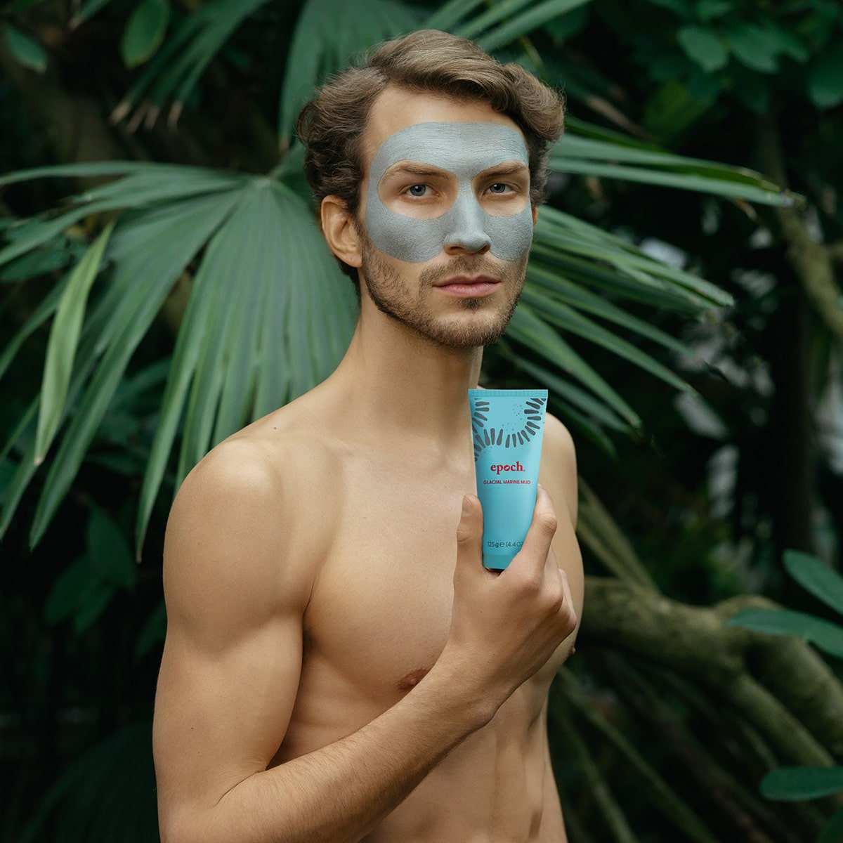 Junger Mann vor dunkelgrünem Farn und trainiertem Oberkörper hält eine Tube des Glacial Marine Mud von Nu Skin. Die Maske ist in seinem Gesicht aufgetragen.